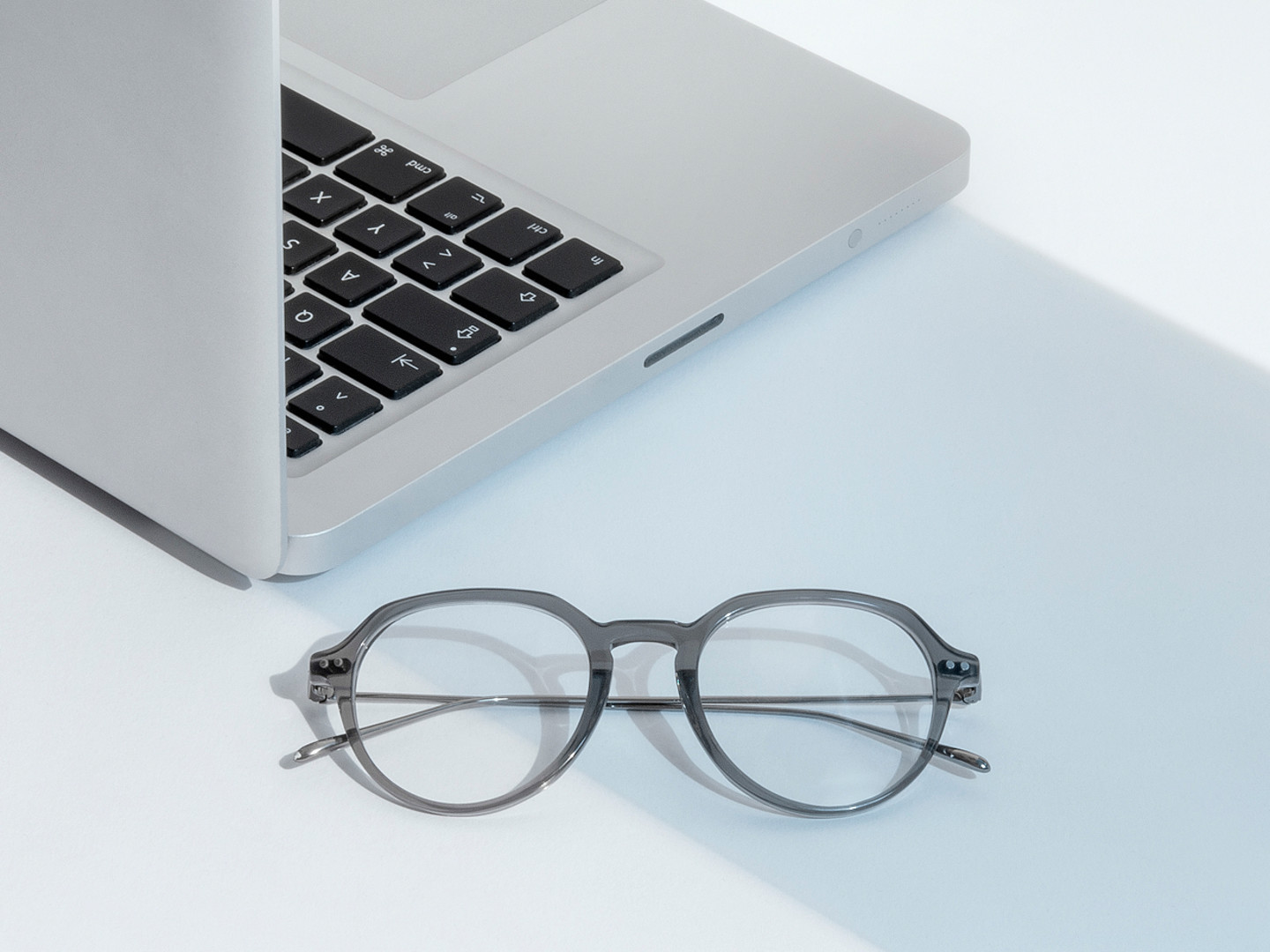 Graue Brille liegt neben einem aufgeklappten Laptop auf hellem Untergrund.